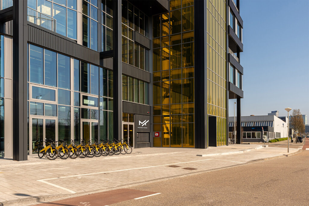 Serviced apartments Entree van de Makerstoren met het logo boven de toegangsdeur, een rij gele deelfietsen langs de glazen gevel en een heldere lucht op de achtergrond.