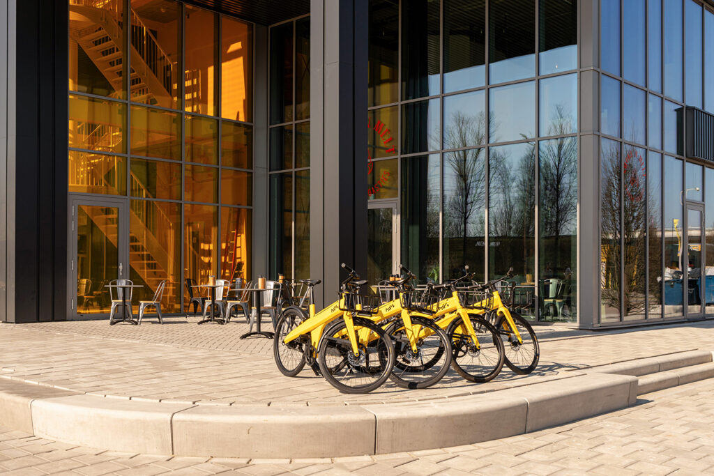 Gele deelfietsen met het Makerstoren-logo geparkeerd voor de glazen gevel van het gebouw, met een terras en warm gouden licht vanuit het interieur op de achtergrond.
