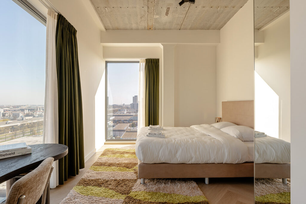 Lichte slaapkamer in de Makerstoren met tweepersoonsbed, groene gordijnen en panoramisch uitzicht over Amsterdam vanuit een hoog gelegen appartement.