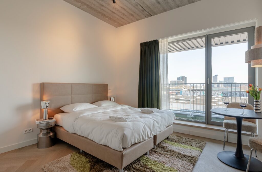 Renting a flat in Amsterdam for 1 month, this is how you do it Bright bedroom of a serviced apartment in Makerstoren Amsterdam with queen-size bed, concrete ceiling and glass balcony door overlooking the city